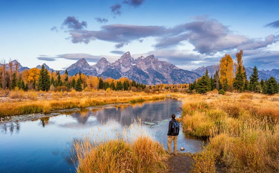 A person standing in front of a lake with trees and mountains in the background.