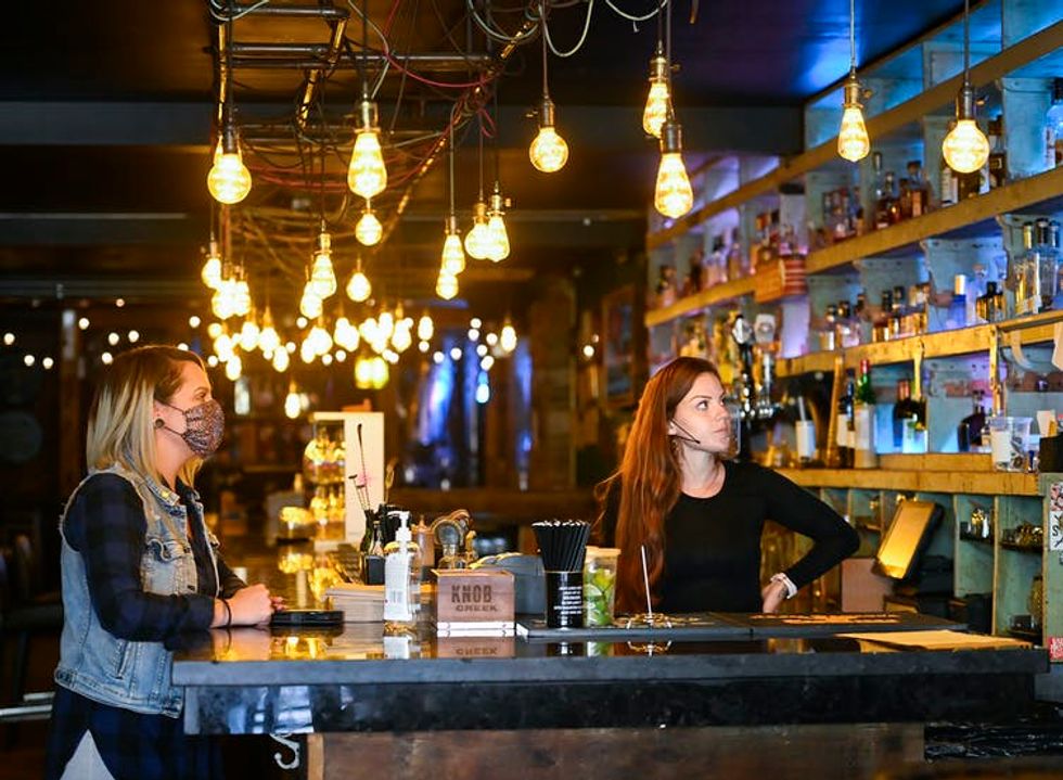 A woman wearing a mask sits at a bar while a bartender stands behind it. Both look at a TV set.